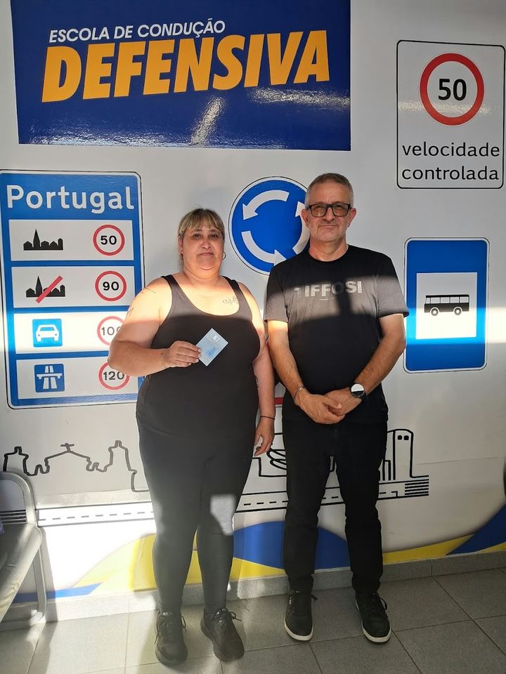 Nova condutora com o instrutor, a mostrar a carta em frente à parede de sinais da escola